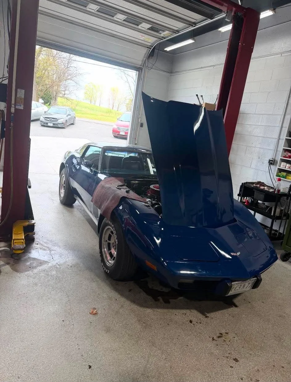Classic C3 Corvette — Dark blue classic C3 Corvette on a two-post lift inside the shop with the hood raised for maintenance.